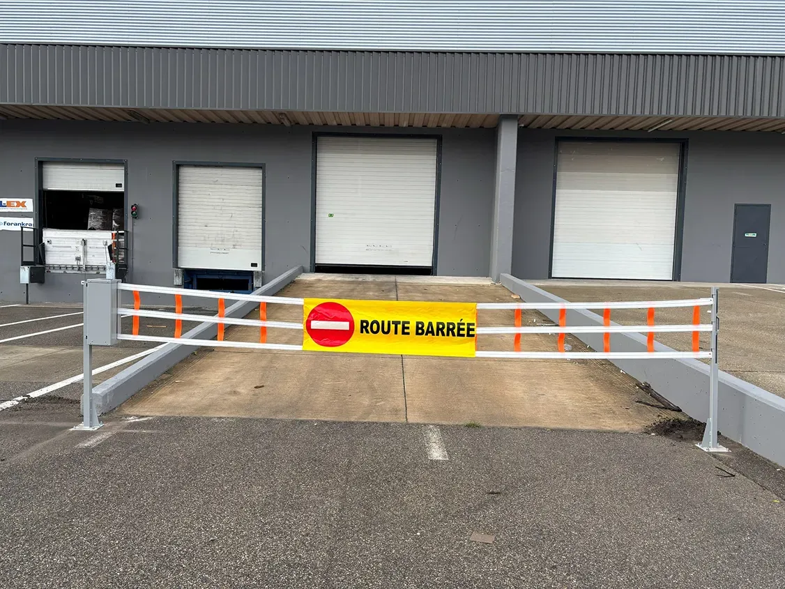 Barrière textile semi-automatique v2 à trois sangles, bannière ROUTE BARRÉE, fermant l'accès aux quais d'un bâtiment logistique